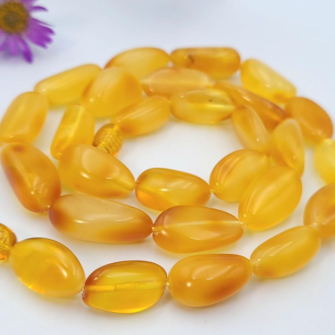 A close-up, high-angle shot of a bright honey-yellow amber necklace laid in a circular pattern on a white surface. The necklace features polished, irregular,
and a purple aster flower is visible in the upper left corner.