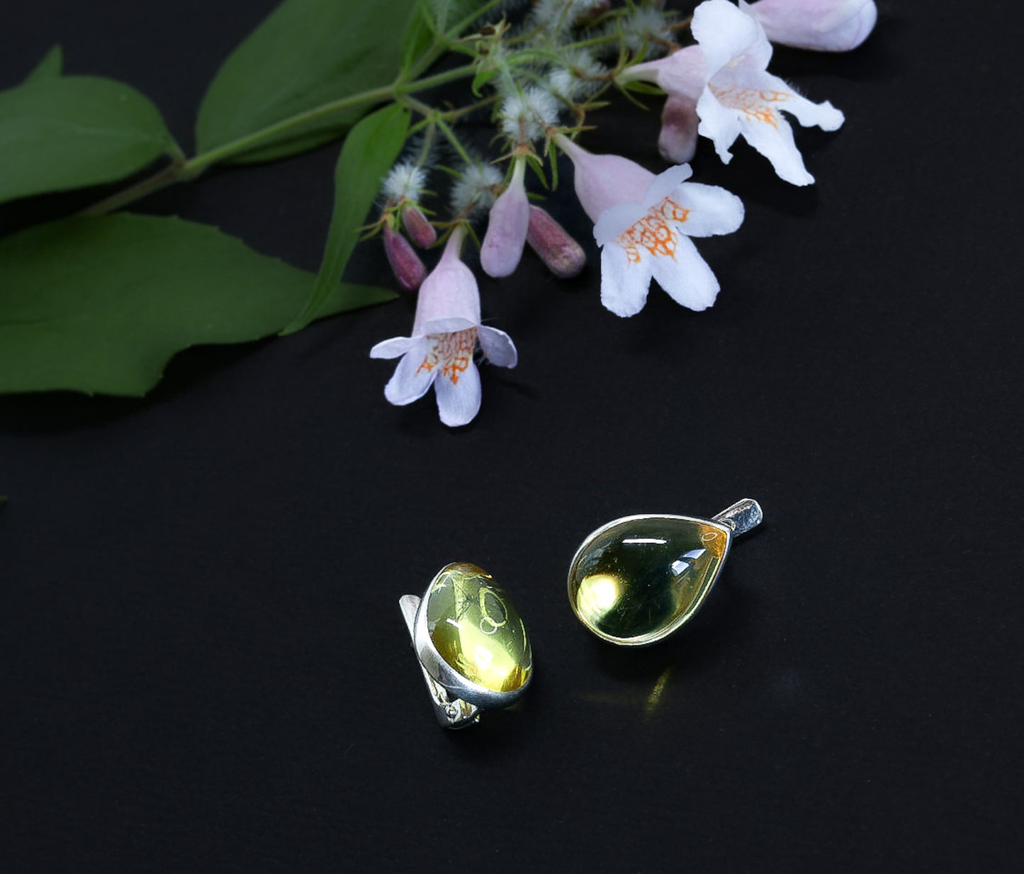 Zwei tropfenförmige Bernstein-Ohrringe mit geschlossener Silberfassung, liegend vor einem Zweig hellrosa Blüten auf schwarzem Untergrund.