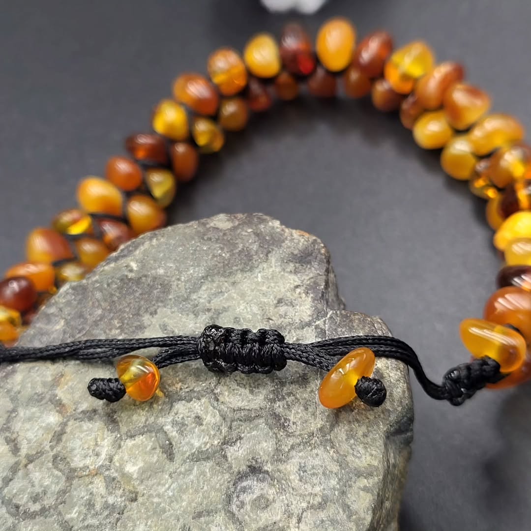 Handgeknüpftes Tetibel Bernstein Armband mit unregelmäßigen, unbehandelten Rohsteinen in Honig- und Cognacfarben, auf einem dunklen Felsen liegend.