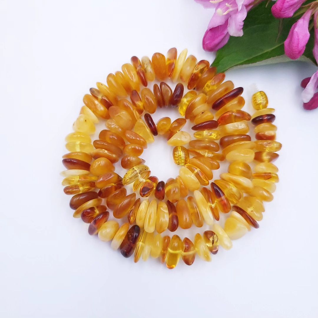 A handmade Tetibel Baltic amber chip necklace coiled in a spiral on a white background with delicate pink flowers.