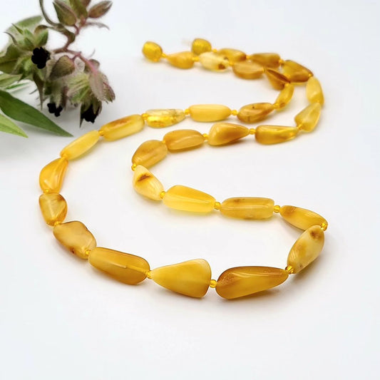 A honey-colored amber teardrop necklace with interspersed small plastic beads, displayed against a white background with a sprig of herbs to the side.