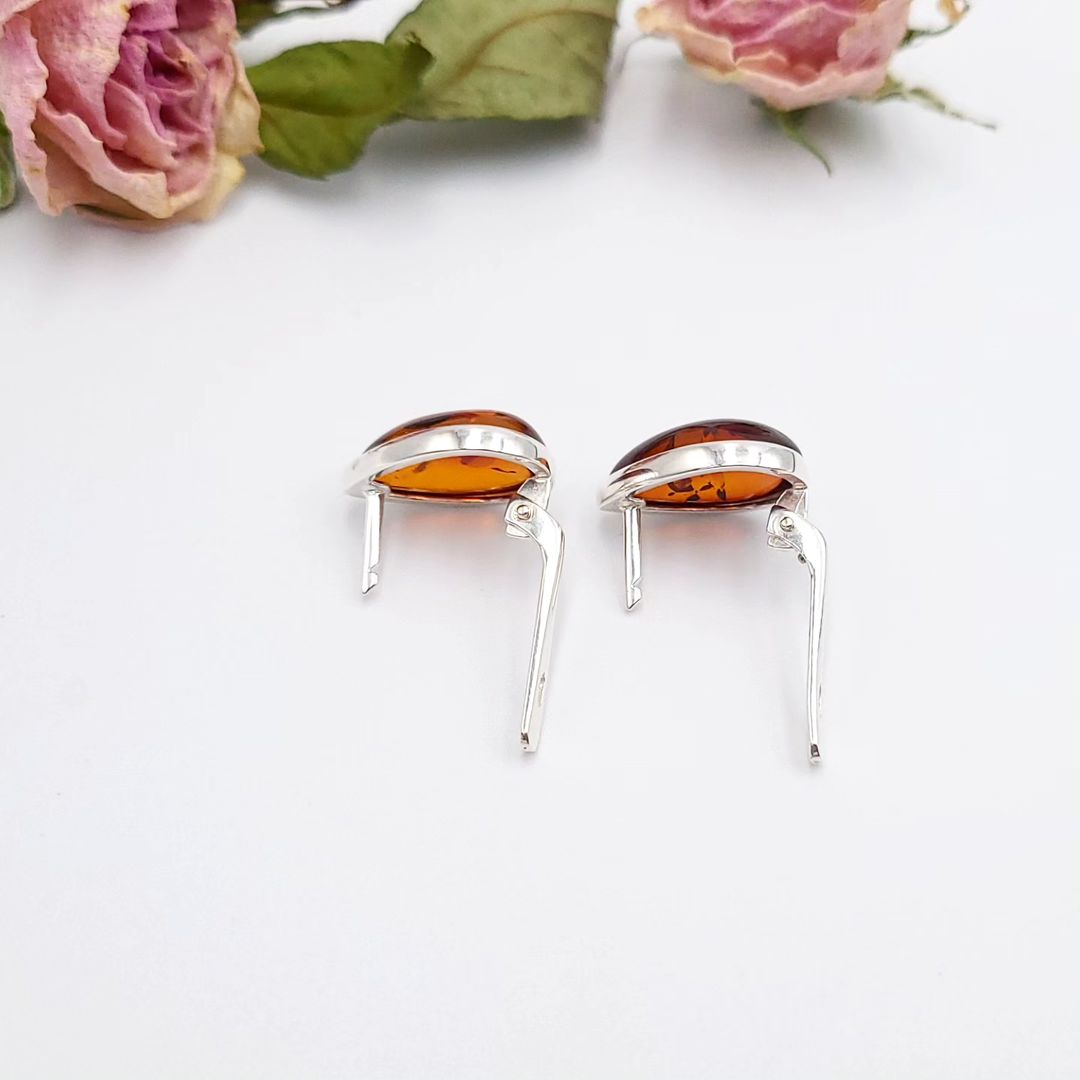 Side view of silver leverback amber earrings, showing the polished silver clasps and the rich, cognac-colored teardrop amber stones, set against a white background with blurred dried pink roses at the top.