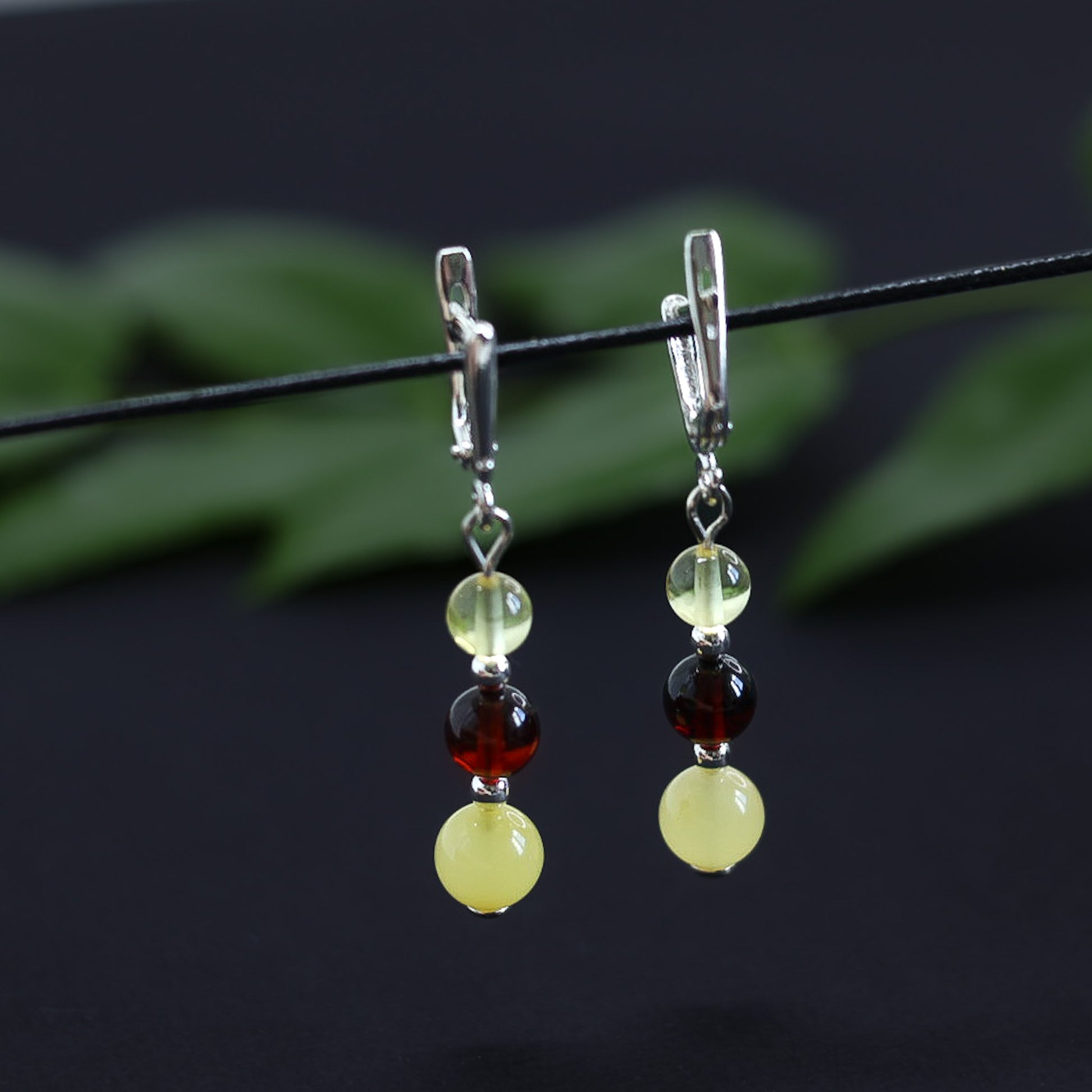 Tetibel Jewels Tricolor Baltische Bernstein Silber Ohrhänger (48mm Länge) hängend vor dunklem, verschwommenem Hintergrund mit grünen Blättern. Die Ohrhänger zeigen Butterscotch, Cognac und Lemon Bernstein Kugeln und glänzende 925 Sterling Silber Haken.