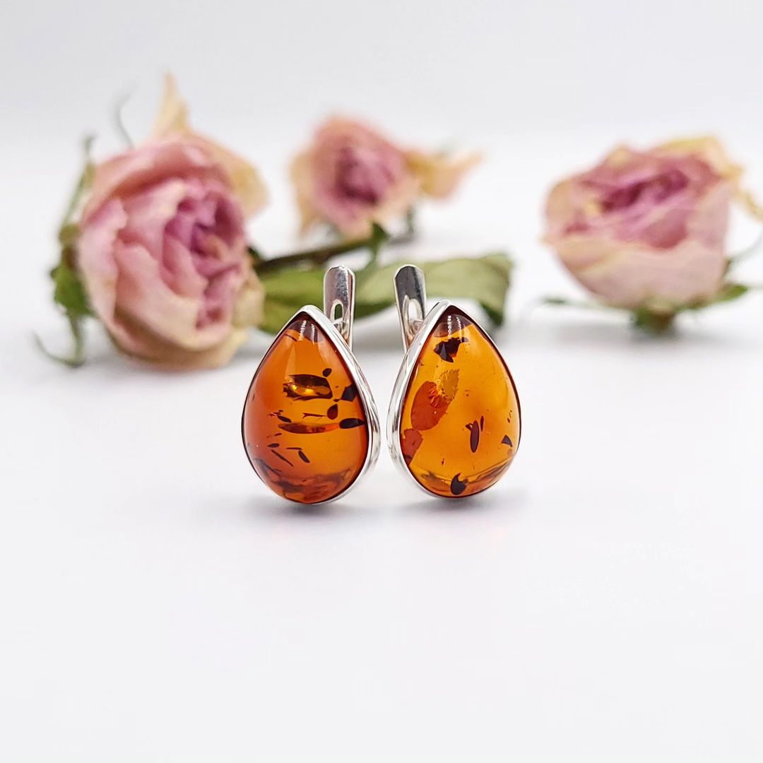 Pair of silver teardrop earrings featuring polished, translucent amber stones with natural dark inclusions, displayed on a white surface with blurred pink dried roses in the background.