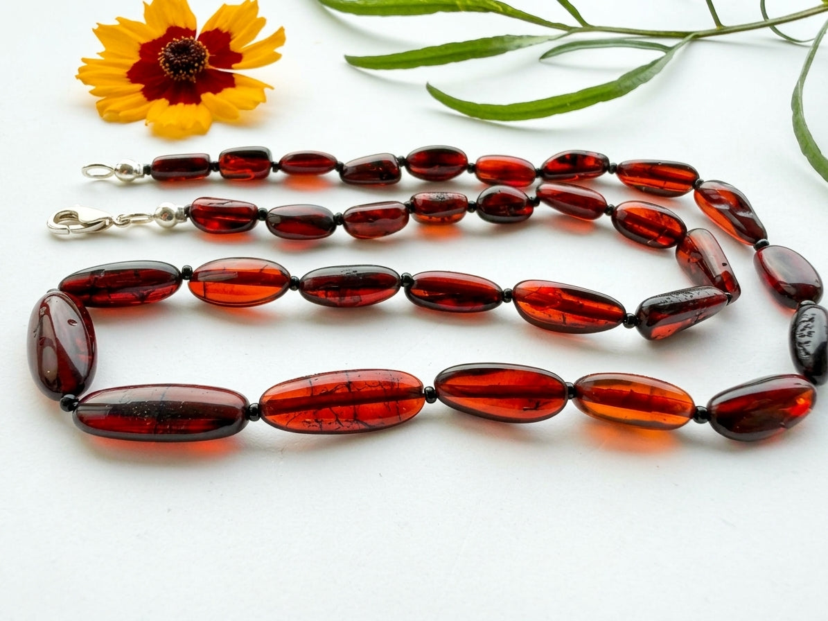 11.6g dark cognac and cherry red Baltic amber bead necklace with protective micro spacer beads lying flat next to a flower.