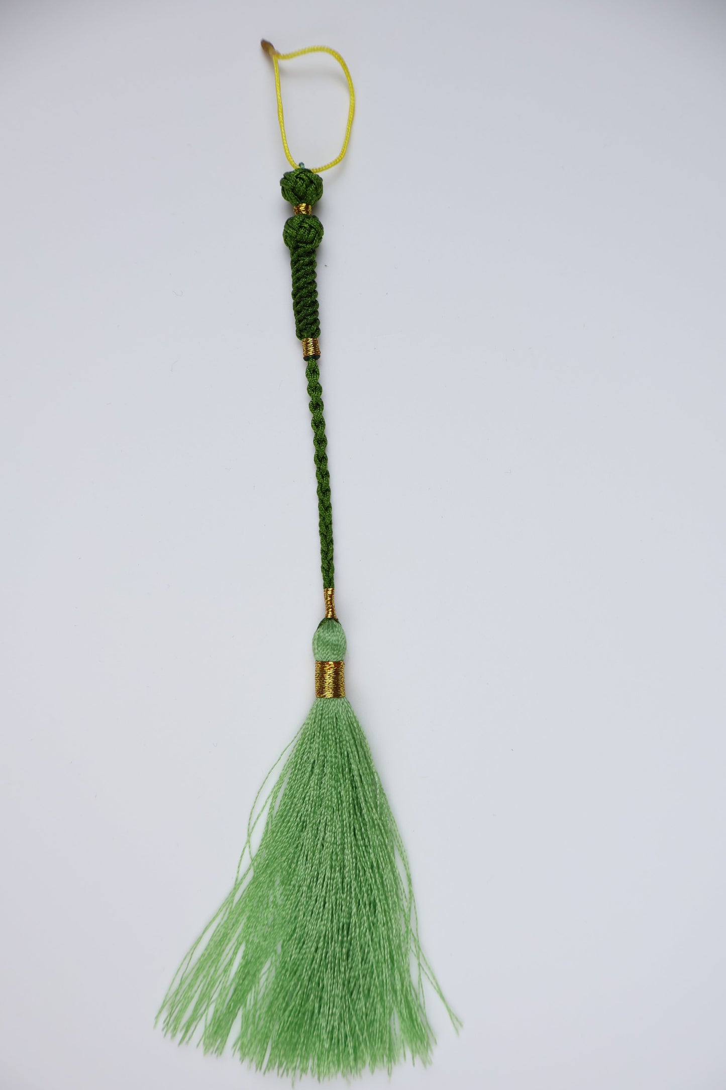 A decorative tassel with a light green silk-like fringe, a dark green braided cord, a small light green bead, and gold-toned metal accents, shown on a plain light gray background.