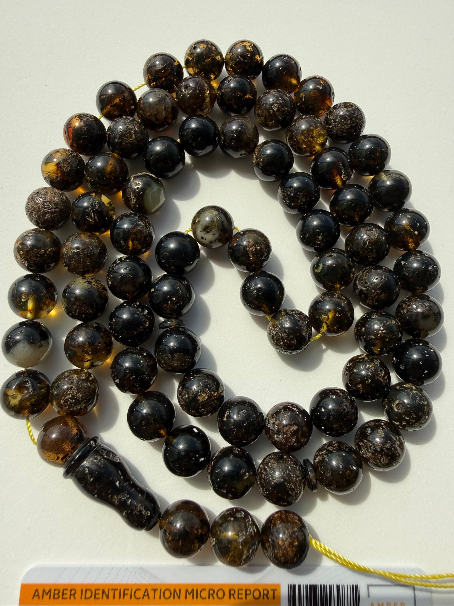 A strand of dark brown and black amber beads with inclusions, coiled on a light background. A carved dark wooden tassel is attached with a yellow string, and a small label at the bottom reads 'AMBER IDENTIFICATION MICRO REPORT'.