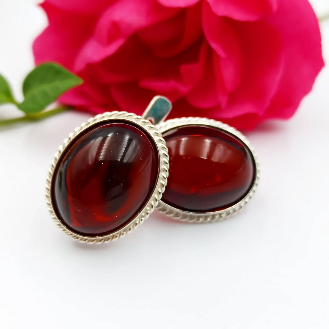 Eleganter, ovaler, kirschroter Bernstein-Ohrschmuck in Silberfassung, präsentiert vor einer pinken Rose.
